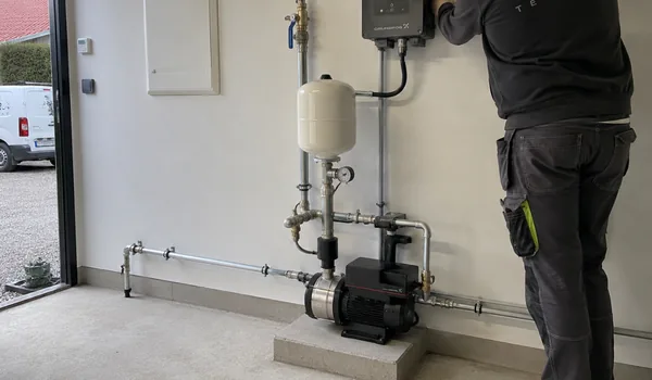 Booster pump on concrete base and white diaphragm tank with pressure gauge, the plumber stands by the control block on the wall, after installation, Tallinn Torutööd shows the customer how to reset the pressure switch.