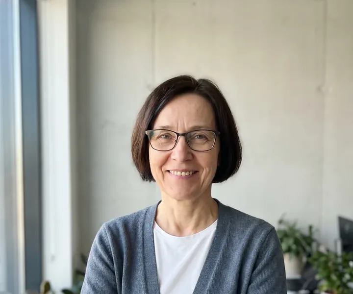 Anneli Kurg tumeda poisipea, prillide ja halli kampsuniga naeratab betoonseina ees akna kõrval, taga toataimed, plaanilise töö kinnituses palub kontor kliendil jätta veemõõtja kapi ette vaba käiguruum.