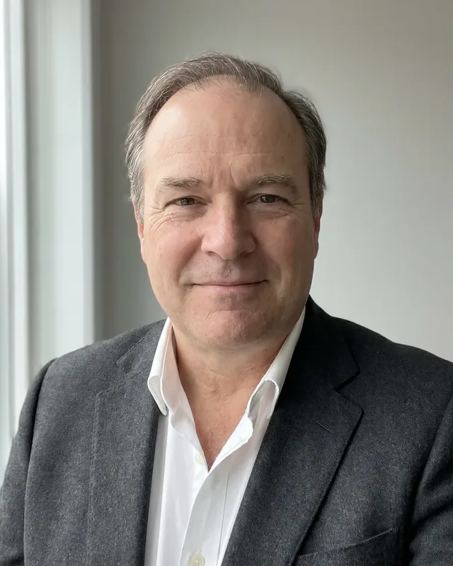 Toomas Kurg with short gray-brown hair, a gray woolen jacket and a white shirt with an open collar looks at the camera in front of a light wall next to the window, before drawing the diagram of the shut-off valves of the contract building, the founder closes and opens each valve once.