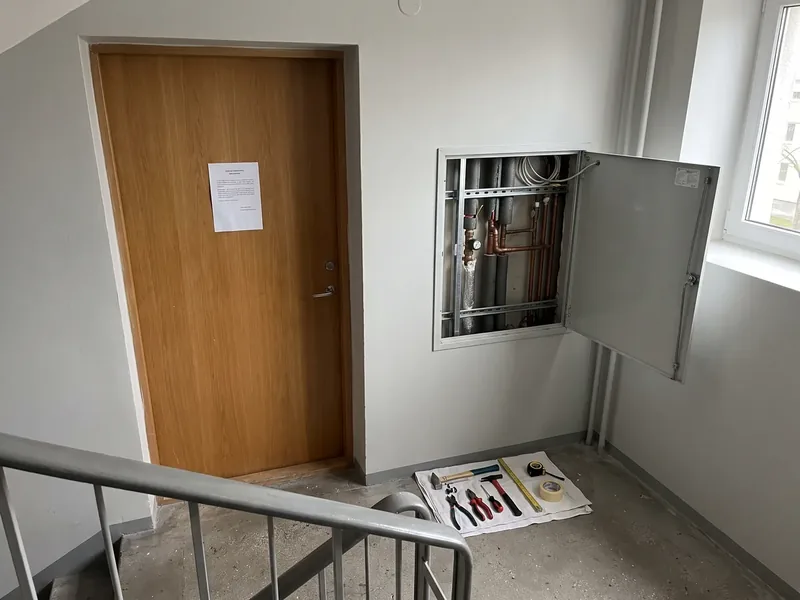 Lasnamäe paneelmaja trepikoda: korteri puituksel prinditud teade, seinas avatud tehnošahti kapp vasktorude ja veemõõtjaga ning põrandal lapil tööriistad, püstiku vahetusel saavad korterite harud tehnošahtis uued messingist kuulkraanid.