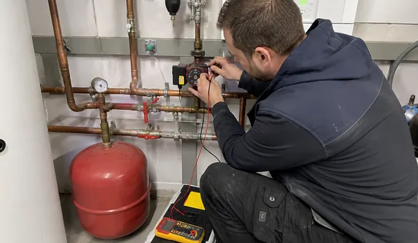 Circulation pump repair, the plumber connects the measuring leads of the multimeter to the pump next to the red expansion tank, in 2025, 71 circulation pumps were replaced in the heating units of the apartment buildings.