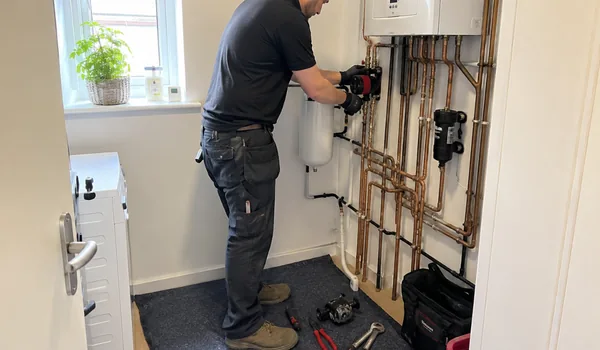 Circulation pump replacement, gloved plumber installs red pump between copper pipes under wall-mounted unit, automatic air separator installed next to pump releases trapped air in circuit before it reaches pump.