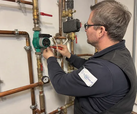 A bespectacled plumber connects the wires of the green circulation pump on the copper piping, a drip loop is left on the pump power cable so that the condensate dripping from the pipe does not run along the cable to the terminal box.